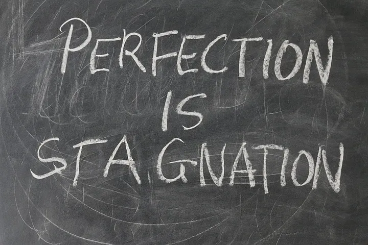 "PERFECTION IS STAGNATION" written in chalk on a chalkboard that has scratches and markings in the background.