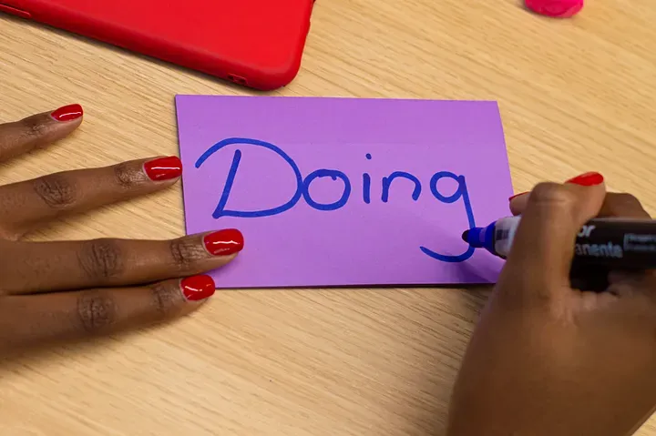 A closeup of a woman's hands writing "Doing" on a purple index card in blue marker.