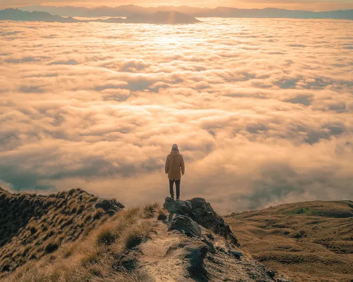 Someone on a mountain peak above the clouds looking at another peak in the distance.